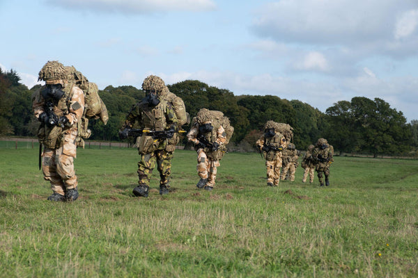 A line of soldiers in camouflage uniforms, gas masks, and helmets covered in netting walks across a grassy field carrying rifles and heavy packs.