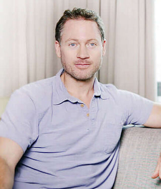 Adult man with short hair sitting on a couch, wearing a purple shirt, looking at the camera.