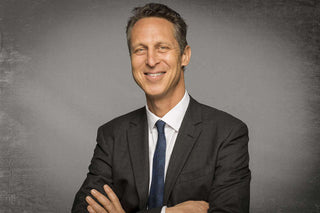 Man in a suit smiling confidently, with arms crossed against a textured gray background.