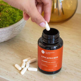 Person taking a capsule from a Neurogan Health Epicatechin bottle with white pills on a marble surface.