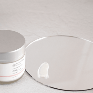 Neurogan GHK-Cu copper peptide cream jar beside a mirror with a dollop of cream on its surface.