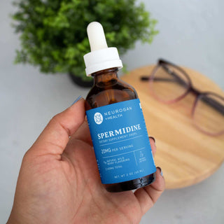 Person holding Neurogan Health Spermidine dietary supplement dropper bottle, with glasses and wooden table background.