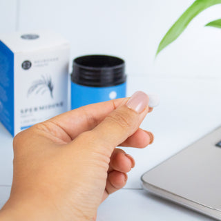 Hand holding a small tablet in front of a skincare product and a laptop, emphasizing wellness and skincare.
