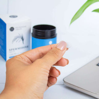 Hand holding a small tablet in front of a skincare product and a laptop, emphasizing wellness and skincare.