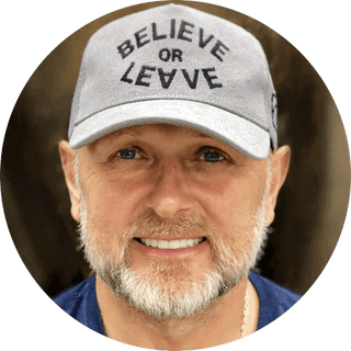 Close-up of a man wearing a cap that says 'BELIEVE OR LEAVE', smiling confidently.