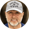 Close-up of a man wearing a cap that says 'BELIEVE OR LEAVE', smiling confidently.