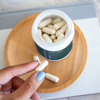 Hand holding a white capsule near an open jar of NAD+ capsules on a wooden plate.