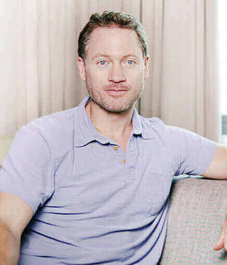 Adult man with short hair sitting on a couch, wearing a purple shirt, looking at the camera.