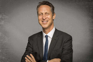 Man in a suit smiling confidently, with arms crossed against a textured gray background.