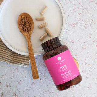 HTB capsules in amber bottle with vegetable capsules spilled beside a wooden spoon filled with buckwheat seeds on a white plate.