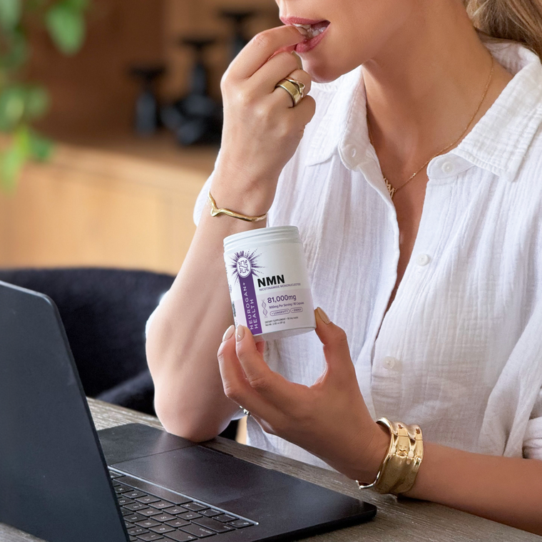 A woman at her laptop taking a capsule, holding the NMN Capsules 900 mg jar