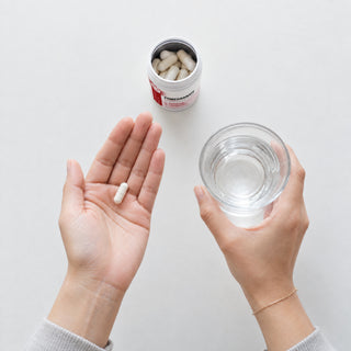 Pomegranate extract capsule in an open palm beside a glass of water, with an open tub of Neurogan Health Pomegranate Extract capsules behind