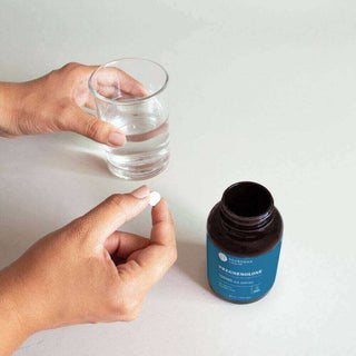 Person holding a white supplement capsule near a glass of water and an open pregnenolone bottle.