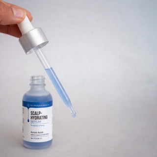 Hand holding a glass dropper with a drop of blue Scalp-Hydrating Serum forming at the tip, positioned above the open bottle on a light grey background.