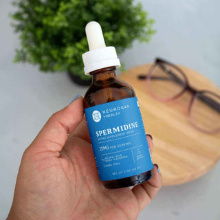 Person holding Neurogan Health Spermidine dietary supplement dropper bottle, with glasses and wooden table background.