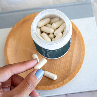 Hand holding a white capsule near an open jar of NAD+ capsules on a wooden plate.