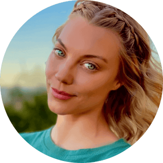 Smiling woman with braided hair and blue eyes outdoors, wearing a teal shirt and surrounded by nature.