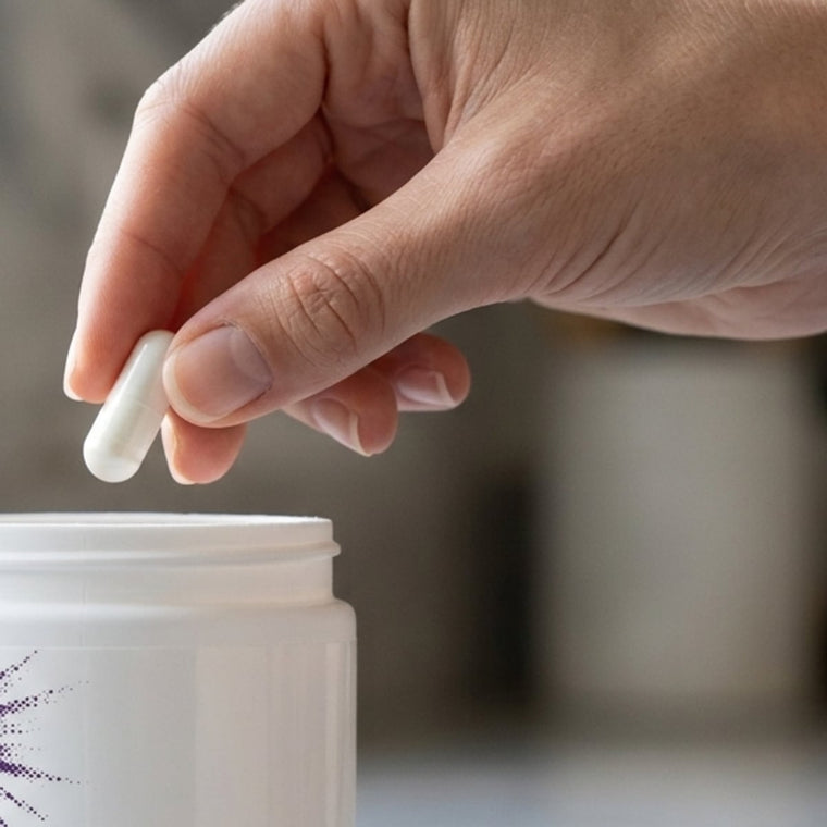 A hand lifting one white capsule out of an open NMN 900 mg jar